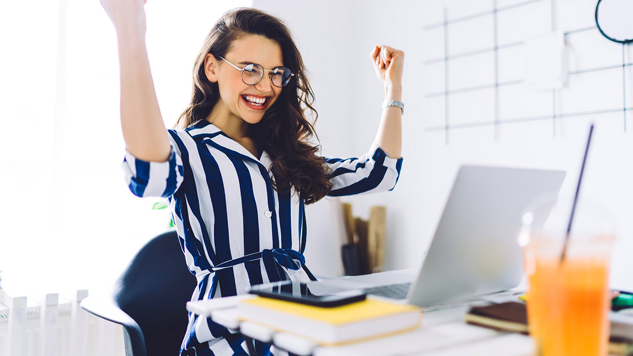 Overjoyed female freelancer in glasses and casual clothes sitting at desk with laptop with raised arms in winner gesture and celebrating achievements
