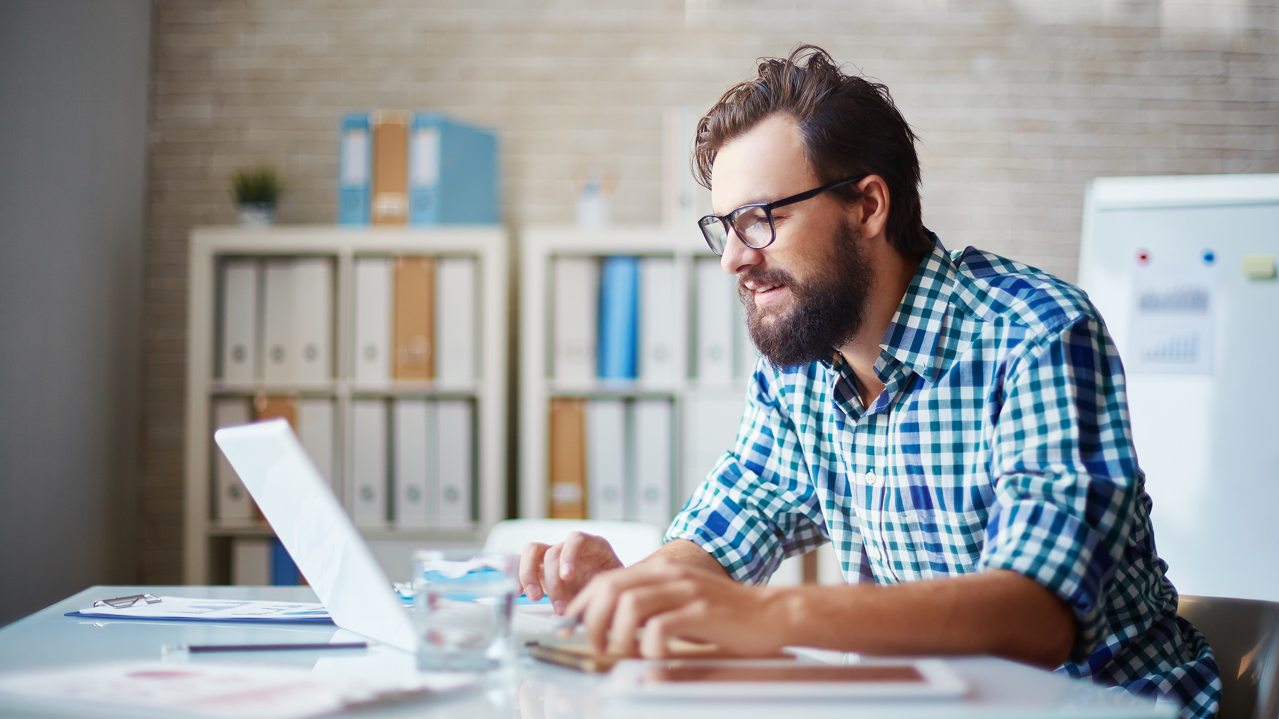 A man sits in front of his laptop at a table, typing on the keyboard and smiling.