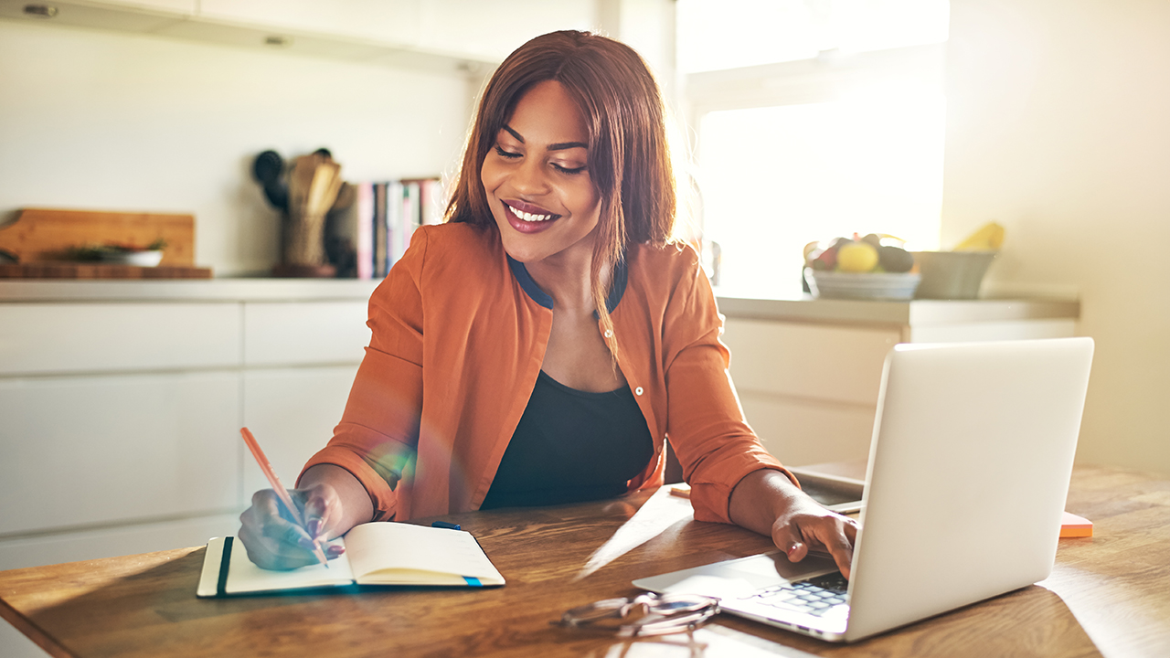 A female coach takes notes while sitting in front of her laptop and studying industry trends in a bright, sunlit room.