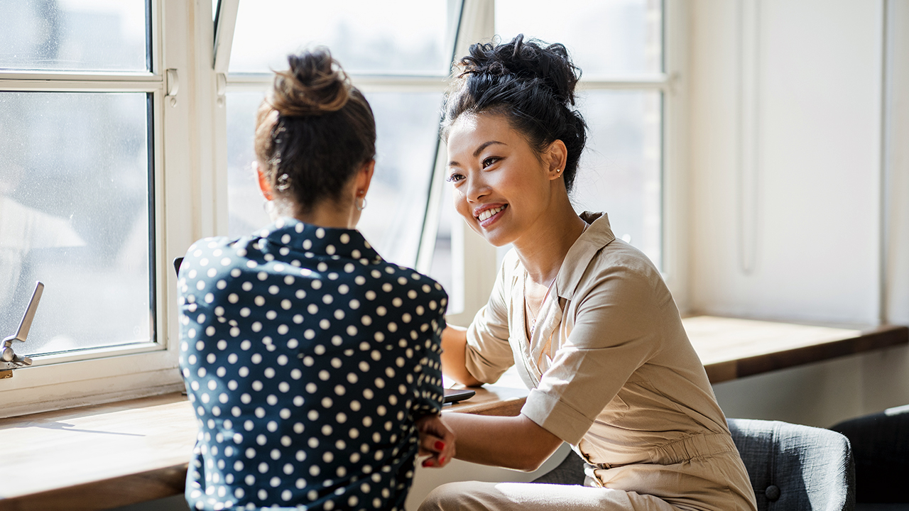 Two women smile at each other while sitting near a window during a coaching session.